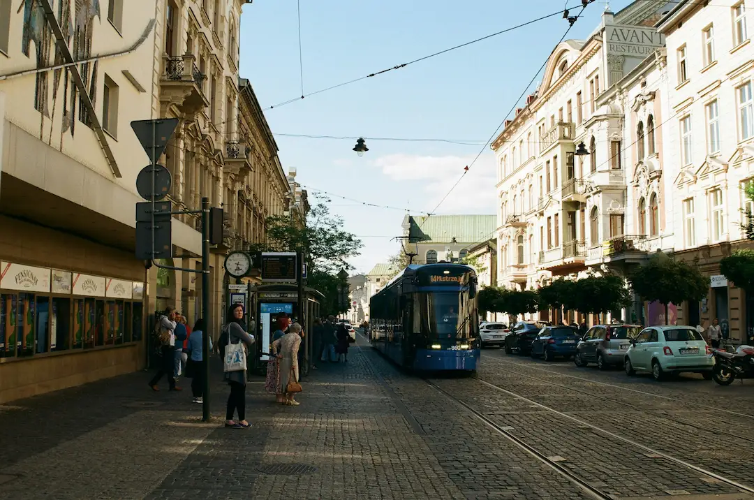 Niebieski tramwaj jedzie po torach ułożonych z bruku historyczną ulicą miasta, w otoczeniu ozdobnych budynków, zaparkowanych samochodów, trakcji elektrycznej i pieszych, skąpanych w ciepłym świetle popołudnia.