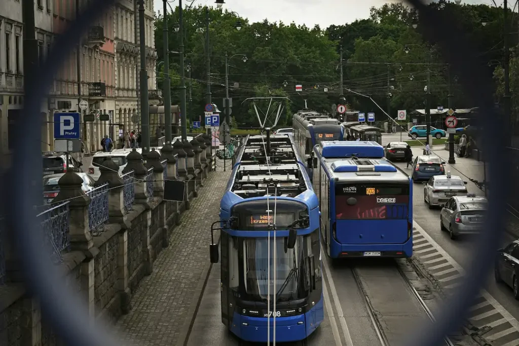 Dwa niebieskie tramwaje poruszają się po torach biegnących obok jezdni na ruchliwej, zadrzewionej ulicy; widok przez ciemne ogrodzenie nadaje miejskiej scenerii przytłumiony, spokojny charakter.