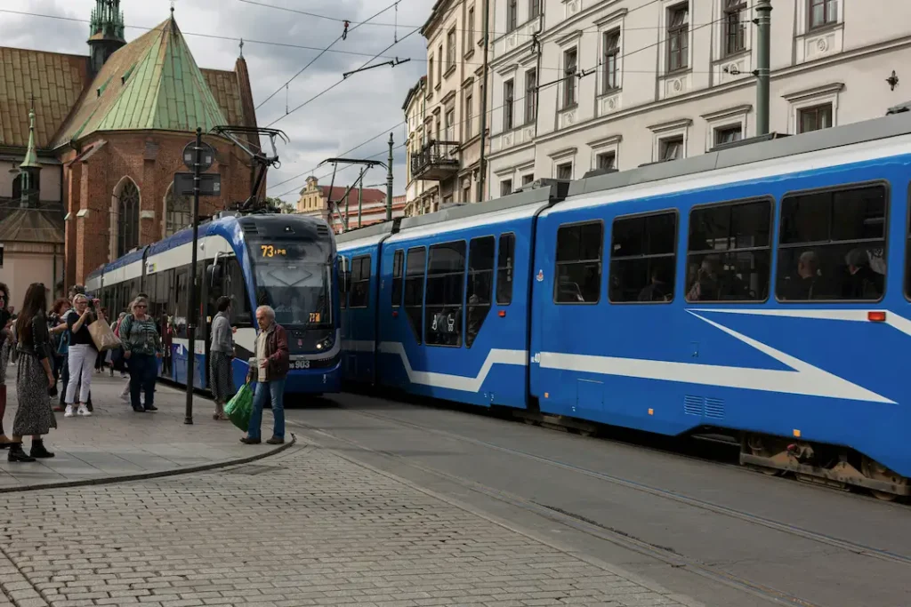 Niebieski tramwaj zatrzymuje się na brukowanym przystanku, do którego wsiadają pasażerowie, na tle zabytkowych europejskich budynków i przewodów napowietrznych; codzienny, tętniący życiem miejski krajobraz.